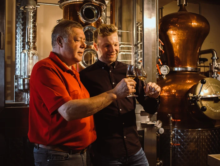 Zwei Männer stehen in einer Brennerei bei glänzenden Kupferdestillen und probieren gemeinsam Spirituosen.