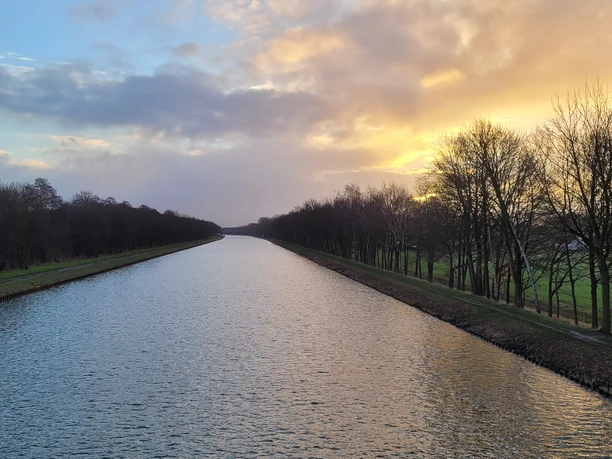 Sonnenuntergang über dem Mittellandkanal, umgeben von Bäumen und Gräsern, sanftes Licht reflektierend.