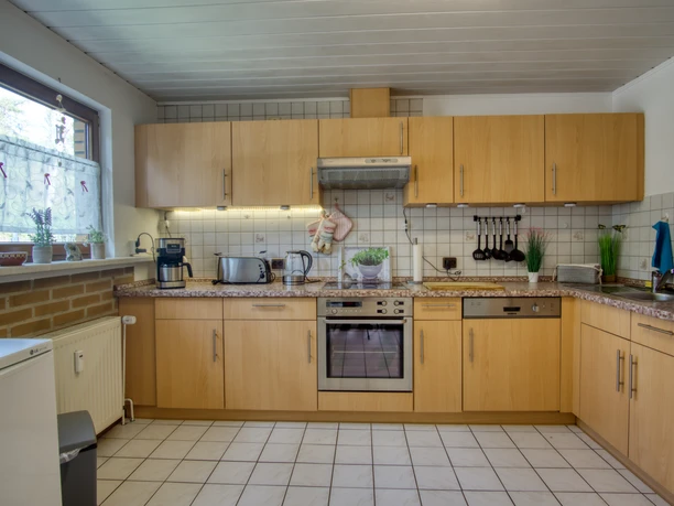 Küche Helle Einbauküche mit Holzfronten, Fliesenboden und Arbeitsplatte in Natursteinoptik. Ausgestattet mit Backofen, Herd, Spülmaschine, Wasserkocher, Toaster und Kaffeemaschine. Ein Fenster bringt Tageslicht, auf der Fensterbank stehen Pflanzen und kleine Dekoelemente.