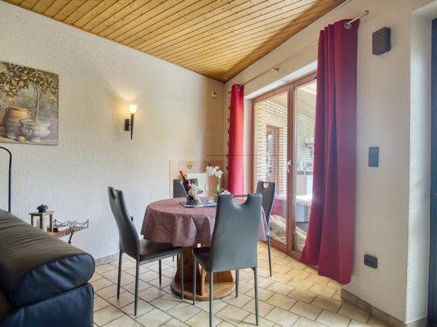 Sitzecke Heller Wohnbereich mit rundem Holztisch und vier schwarzen Stühlen vor einer großen Fensterfront mit roten Vorhängen. Eine schwarze Ledercouch steht seitlich, an der Wand hängt ein Stillleben mit Gefäßen. Decke aus Holzpaneelen und gefliester Boden sorgen für eine warme Atmosphäre.