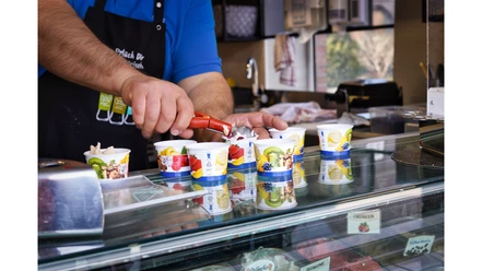Ein Mitarbeiter portioniert mit einem roten Portionierer Eis in bunte Pappbecher auf der Theke.