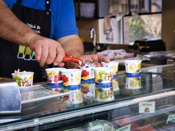 eis-amelinghausen-2022 Ein Mitarbeiter portioniert mit einem roten Portionierer Eis in bunte Pappbecher auf der Theke.