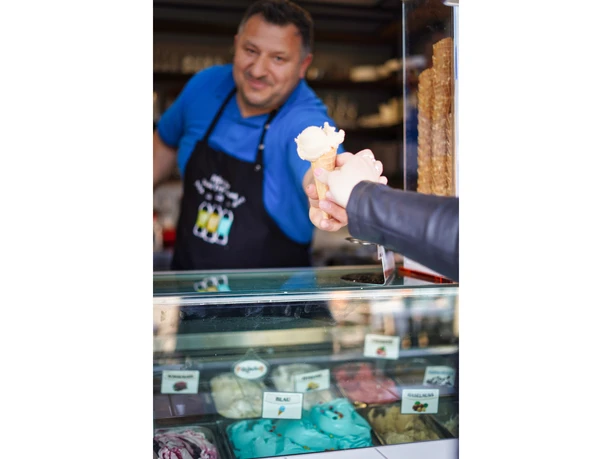 eis-amelinghausen-2022-7.jpg Eisverkäufer reicht einer Kundin lächelnd eine Waffel mit einer Kugel hellem Eis über die Theke.