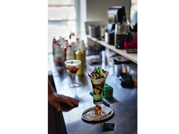 eis-amelinghausen-2022.jpg Hoher Eisbecher mit grünem Eis, Sahne und Schokosoße auf einer Theke mit Arbeitsutensilien.