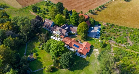 Haus_Berkenkamp.jpg Luftaufnahme eines großen Anwesens mit roten Dächern, umgeben von üppigem Grün und anliegenden Feldern.