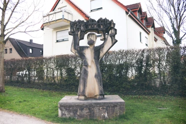 IMG_0248.JPG Steinskulptur eines Mannes mit erhobenen Armen, der ein detailreiches Modell einer Stadt hält.