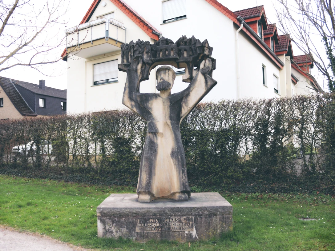 IMG_0248.JPG Steinskulptur eines Mannes mit erhobenen Armen, der ein detailreiches Modell einer Stadt hält.
