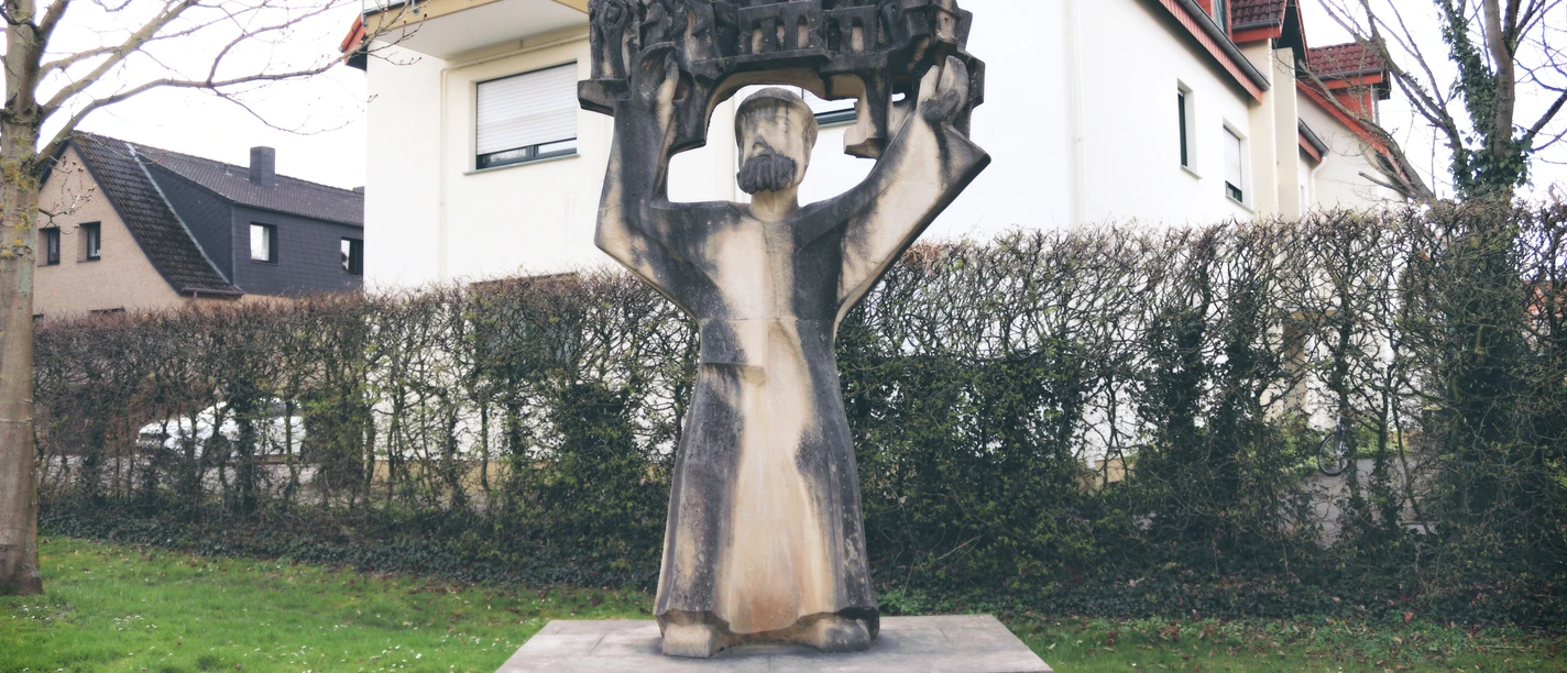 IMG_0248.JPG Steinskulptur eines Mannes mit erhobenen Armen, der ein detailreiches Modell einer Stadt hält.