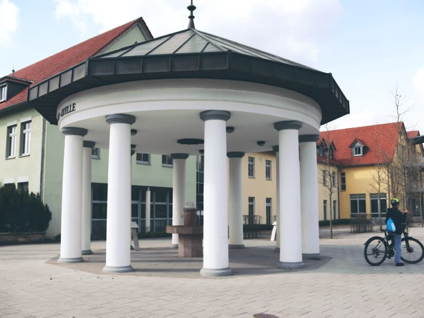 Brunnen mit Säulen und achteckigem Dach auf einem gepflasterten Platz, Radfahrer betrachtet die Szene.