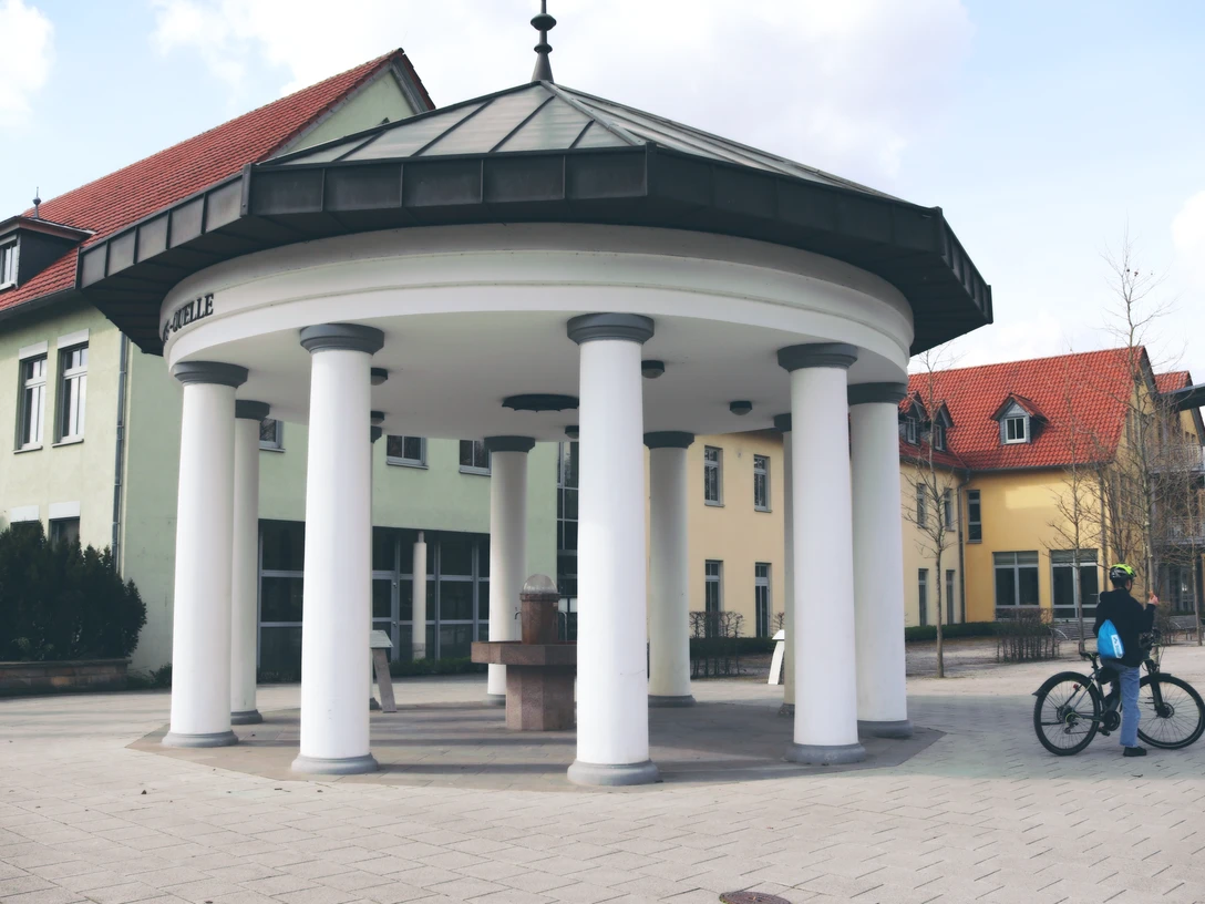 IMG_0183.JPG Brunnen mit Säulen und achteckigem Dach auf einem gepflasterten Platz, Radfahrer betrachtet die Szene.
