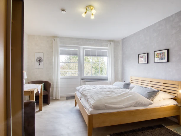 Schlafzimmer Helles Schlafzimmer mit Holzdoppelbett, grauer Wand, Sitzecke und großem Fenster.