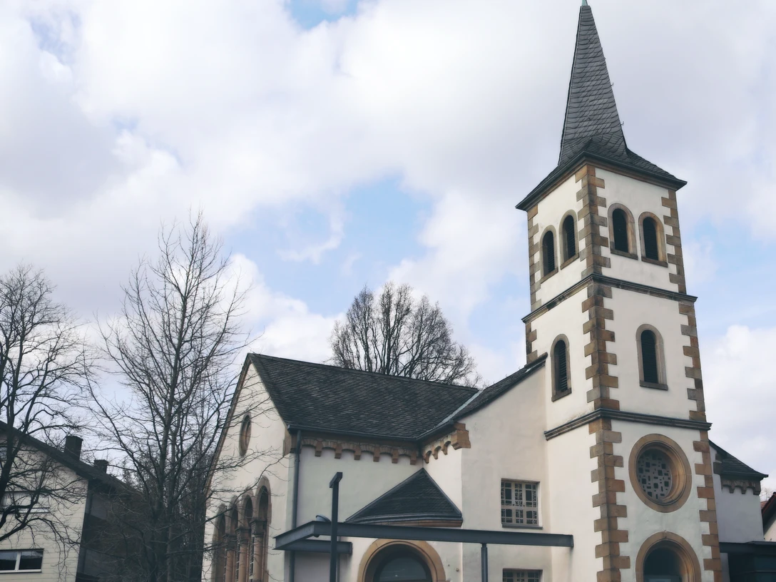 Kirche mit hohem Turm, Wetterfahne, umgeben von Bäumen und Autos; klarer Himmel mit einigen Wolken.