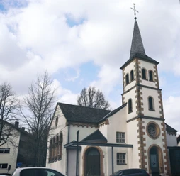IMG_0101.JPG Kirche mit hohem Turm, Wetterfahne, umgeben von Bäumen und Autos; klarer Himmel mit einigen Wolken.