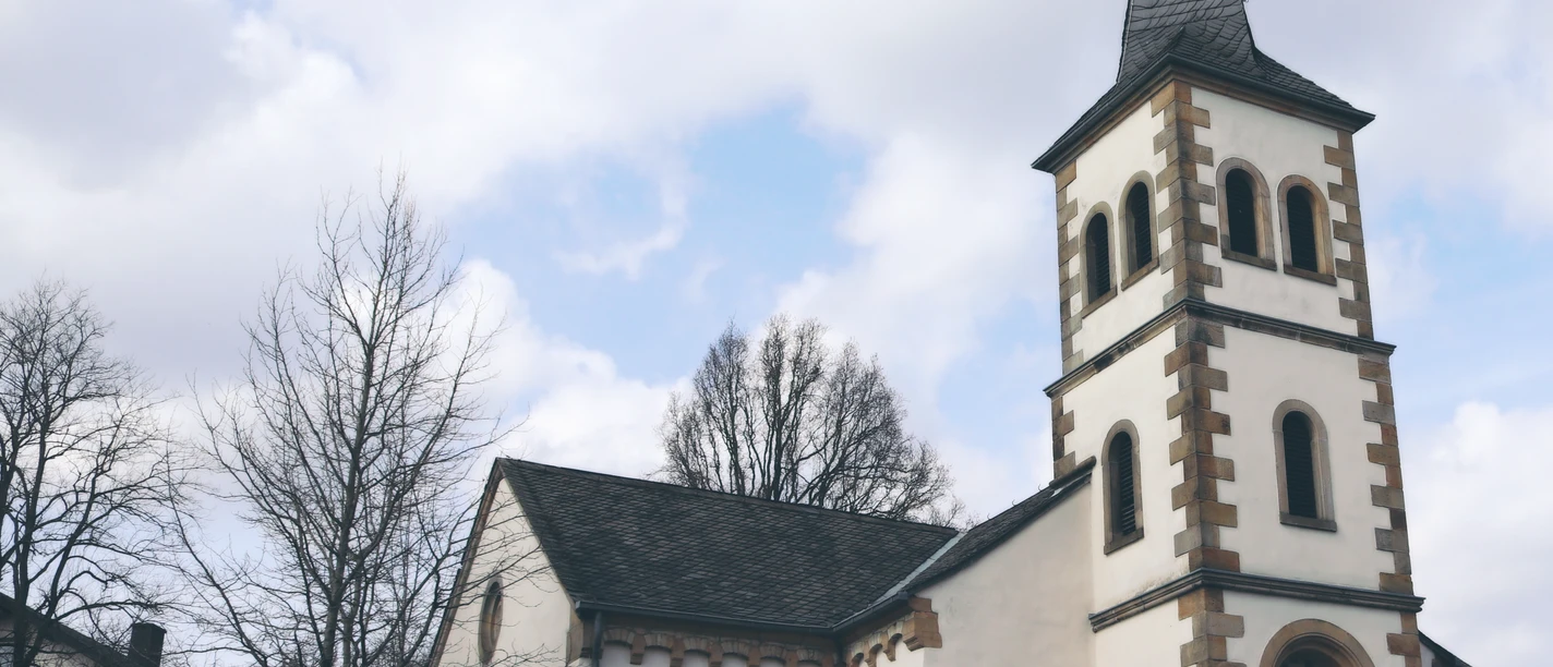 IMG_0101.JPG Kirche mit hohem Turm, Wetterfahne, umgeben von Bäumen und Autos; klarer Himmel mit einigen Wolken.