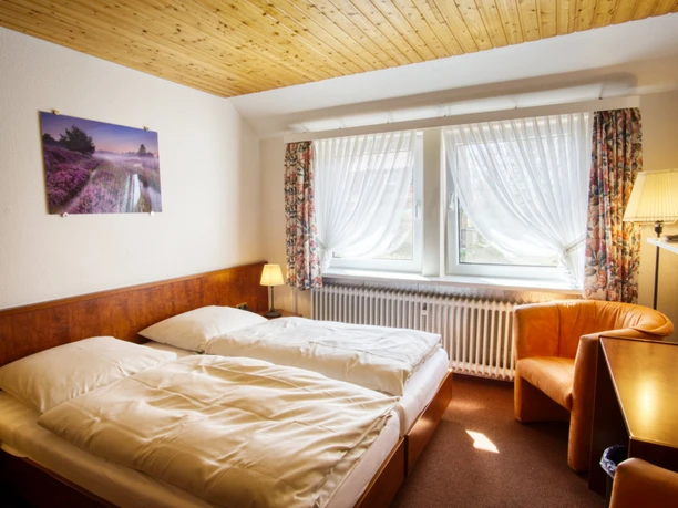 Doppelzimmer Helles Doppelzimmer mit Doppelbett, Holzdecke, orangem Sessel, Fenstern und einem Heidebild.