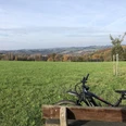 "Aus dem Tal der Agger zum Dom auf der Höhe" Weitblick über eine grüne Landschaft mit sanften Hügeln, Fahrrad an einer Parkbank im Vordergrund.