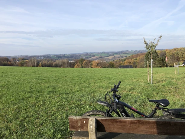 "Aus dem Tal der Agger zum Dom auf der Höhe" Weitblick über eine grüne Landschaft mit sanften Hügeln, Fahrrad an einer Parkbank im Vordergrund.