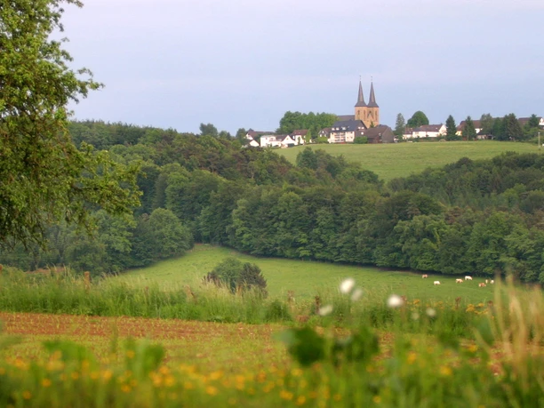 Aus dem Tal der Agger zum Dom auf der Höhe Kirche auf Hügel, umgeben von grünen Wiesen und Wäldern, mit Bäumen im Vordergrund und Dorf im Hintergrund.