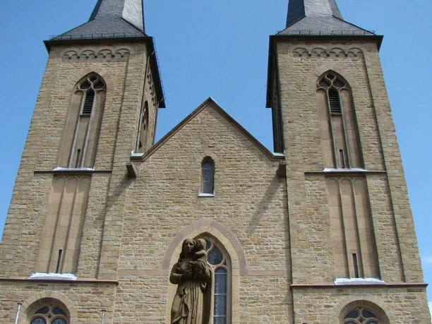 Historische Kirche mit zwei hohen Türmen und einer Heiligenstatue im Vordergrund. Klarer Himmel.