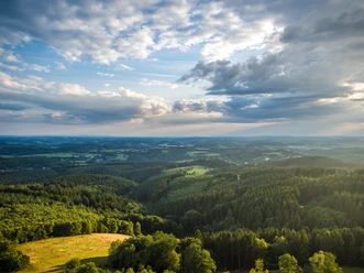 Reichshof-Blockhaus Weite, grüne Landschaft mit dichten Wäldern und Feldern unter einem Himmel mit dramatischen Wolkenformationen.
