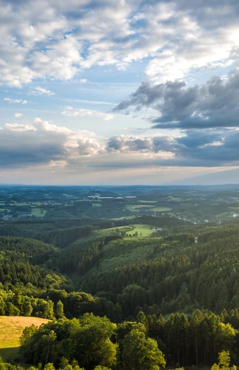 Reichshof-Blockhaus Weite, grüne Landschaft mit dichten Wäldern und Feldern unter einem Himmel mit dramatischen Wolkenformationen.