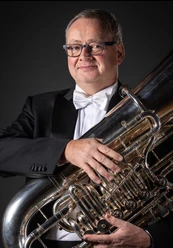 Ein Mann in Anzug hält eine Tuba. Er lächelt und trägt eine schwarze Brille und eine Fliege.