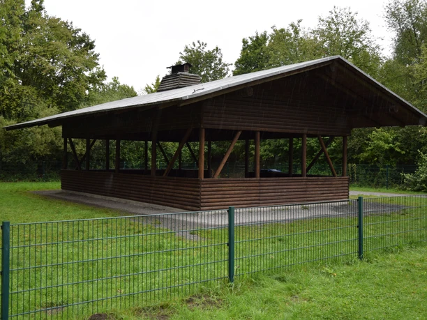 Überdachter Pavillon aus Holz mit offenen Seiten, umgeben von Wiese und Bäumen, fernab urbaner Gebiete.