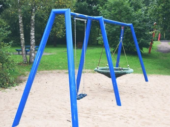 DSC_0083.JPG Blaue Doppelschaukel auf einem Sandspielplatz, umgeben von Bäumen und grüner Vegetation.