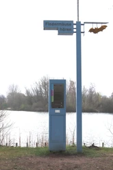 Blauer Audioguide-Pfahl mit "Fledermäuse hören"-Schild vor einem See, umgeben von Bäumen.