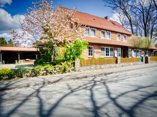 Landgasthaus Eichenkrug Backsteingebäude mit rotem Ziegeldach und Holzverkleidung im Obergeschoss hinter einem blühenden Magnolienbaum.
