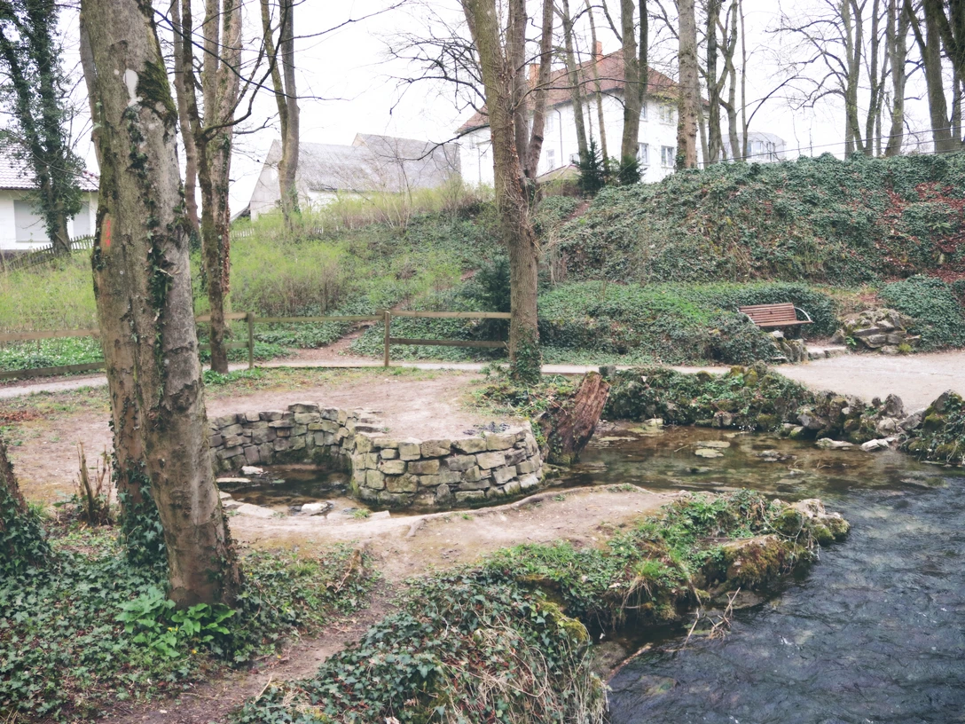 Ein kleiner Brunnen aus Stein steht in einem bewaldeten Park. Ein Bach fließt nebenan vorbei.