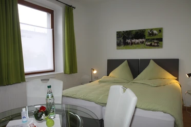 Helles Schlafzimmer mit grünem Doppelbett, Lampe und Tisch mit Wasserflasche und Apfel.