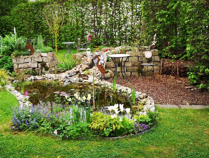 Ein kleiner Gartenteich mit blühender Pflanzenvielfalt, umgeben von grünen Sträuchern und Sitzmöglichkeiten.
