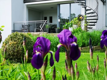 Modernes weißes Haus mit Terrasse und Wendeltreppe, umgeben von einem grünen Garten mit lila Blumen.