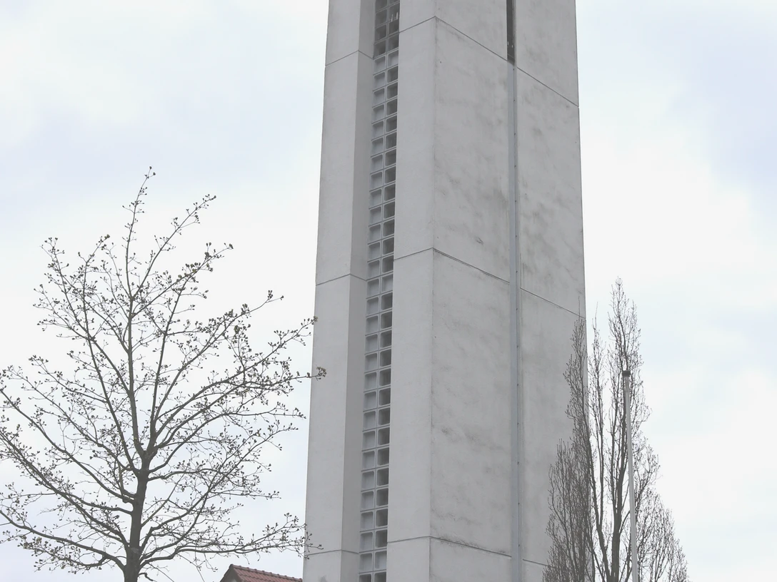 Höher Kirchturm aus Beton neben Wohnhäusern, ein Kreuz an der Spitze und Bäume im Vordergrund.