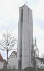 IMG_0563.JPG Höher Kirchturm aus Beton neben Wohnhäusern, ein Kreuz an der Spitze und Bäume im Vordergrund.