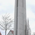 IMG_0563.JPG Höher Kirchturm aus Beton neben Wohnhäusern, ein Kreuz an der Spitze und Bäume im Vordergrund.