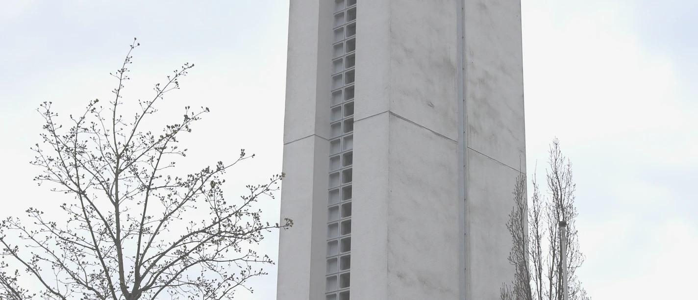 IMG_0563.JPG Höher Kirchturm aus Beton neben Wohnhäusern, ein Kreuz an der Spitze und Bäume im Vordergrund.