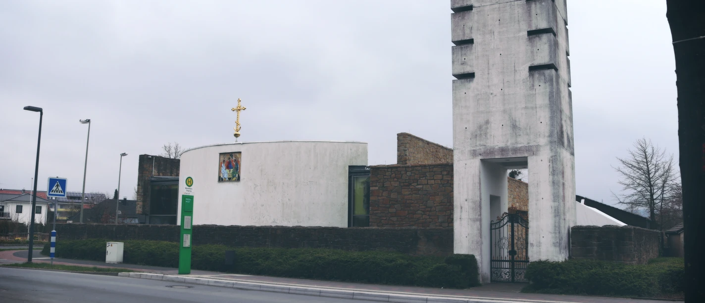 Kirche mit modernem Turm, umgeben von Straßen und Bäumen unter grauem Himmel. Ruhige Umgebung im Bild.