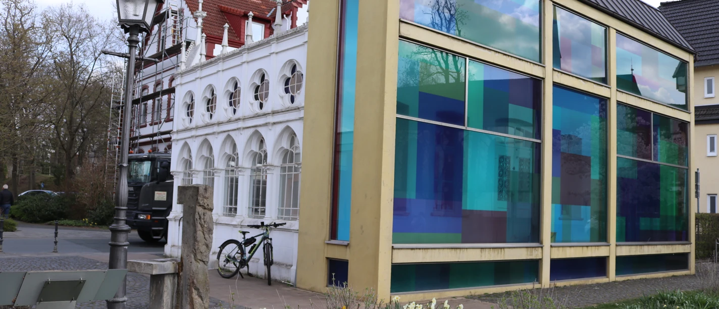 Städtische Ansicht mit modernem Gebäudeteil, bunten Glasfassaden und einer Laterne im Vordergrund.