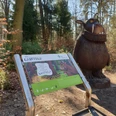 Grüffelo.jpg Grüffelo-Statue im Wald neben einer Informationstafel, umgeben von Bäumen und Laub am Boden.