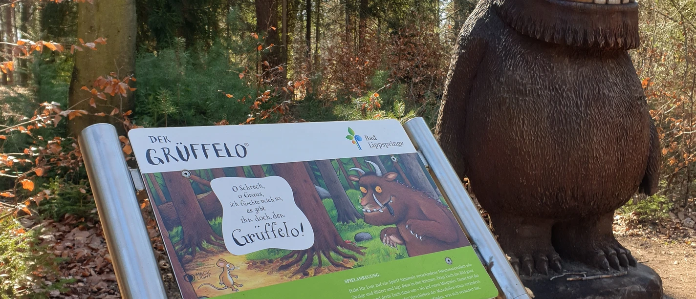 Grüffelo.jpg Grüffelo-Statue im Wald neben einer Informationstafel, umgeben von Bäumen und Laub am Boden.