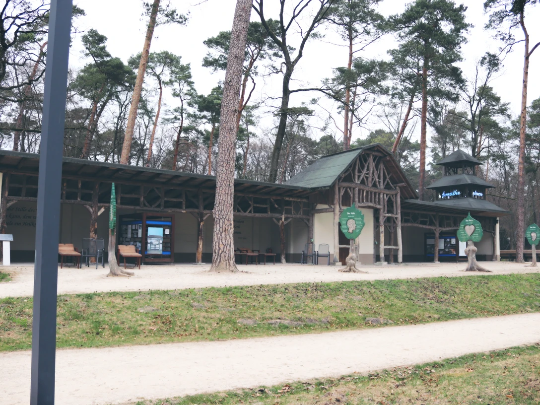 Holzstruktur eines historischen Gebäudes im Wald, umgeben von hohen Kiefern, offener Innenhof.