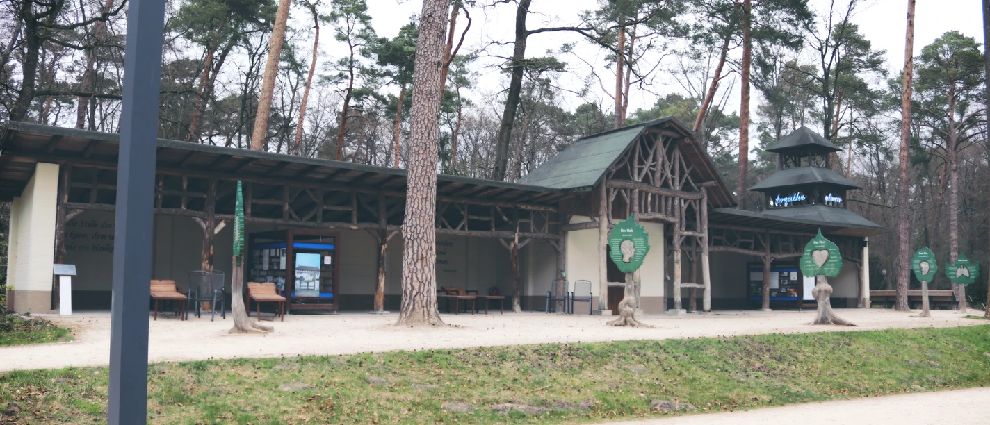 IMG_0572.JPG Holzstruktur eines historischen Gebäudes im Wald, umgeben von hohen Kiefern, offener Innenhof.