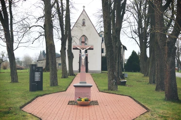 Kreuzweg mit Jesusfigur auf rotem Sockel, flankiert von hohen Bäumen, führt zur Kapelle im Hintergrund.