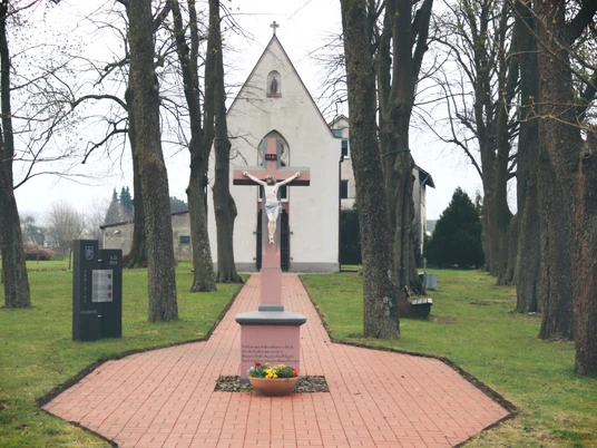 Kreuzweg mit Jesusfigur auf rotem Sockel, flankiert von hohen Bäumen, führt zur Kapelle im Hintergrund.