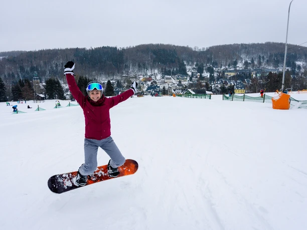 Erlebnisberg Altenberg - Skigebiet Person springt auf Snowboard vor verschneitem Bergpanorama im Skigebiet Altenberg.