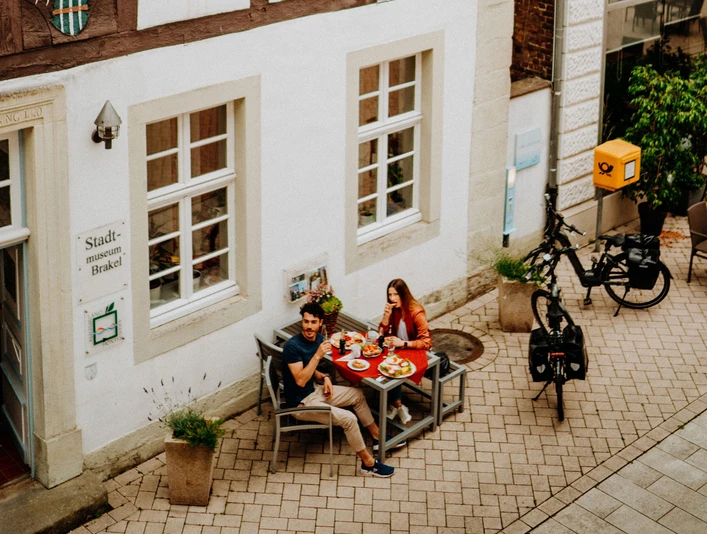 Teutoburger-Wald-Brakel-Tourist-Info-┬®-Teutoburger-Wald-Tourismus-(3).jpg Zwei Personen genießen ein Essen im Freien vor einem historischen Fachwerkgebäude in Brakel.
