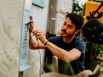 Ein Mann lädt sein E-Bike an einer öffentlichen Ladestation im Freien auf, umgeben von Pflanzen.