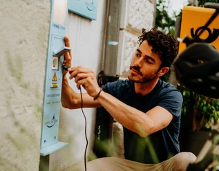 E-Bike Aufladestation Ein Mann lädt sein E-Bike an einer öffentlichen Ladestation im Freien auf, umgeben von Pflanzen.